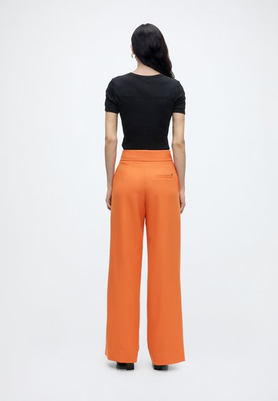 Femme aux longs cheveux foncés portant un haut noir à manches courtes et un pantalon orange taille haute à jambes larges, debout de dos.
