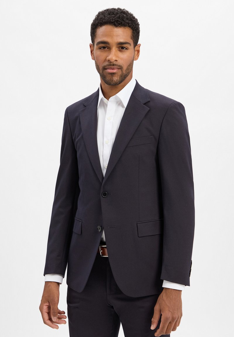 Marineblauwe op maat gemaakte colbert met notch-revers, enkele knoopsluiting en twee voorzakken; gedragen over een wit overhemd.