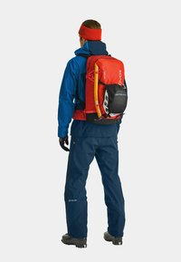 Roter Ortovox-Rucksack mit Ausrüstungsbefestigungen, getragen über einer blauen Jacke und navyfarbenen Hosen. Schwarze Handschuhe und ein Winter-Stirnband runden das Outfit ab.