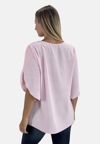 Lichtroze top met losse vleermuismouwen en een ronde halslijn. Zachte textuur, langere achterkant. Gedragen met donkere denim jeans.