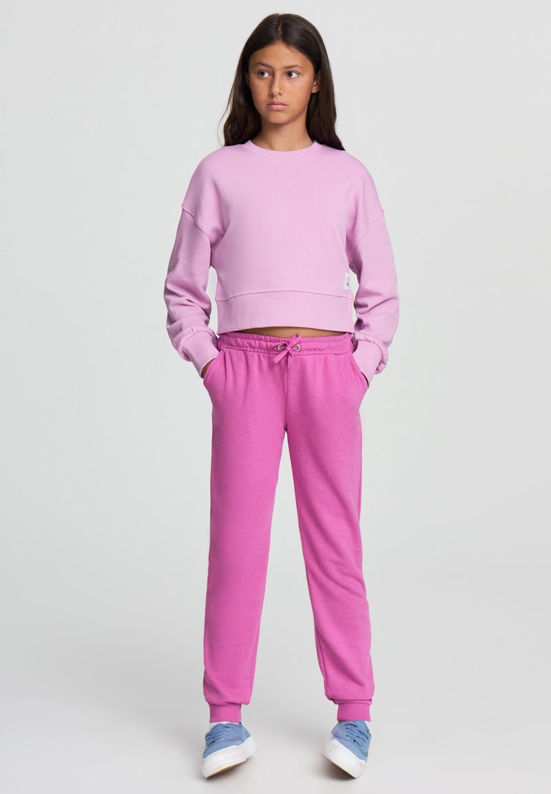 Jasnoróżowa, skrócona bluza oraz dopasowane różowe dresy wykonane z miękkiej bawełny, z elastycznym paskiem w talii i ściągaczami na kostkach. Styl casual.