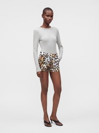 Femme portant un haut gris clair à manches longues, un short imprimé animalier et des sandales noires à talons, debout les mains dans les poches sur fond blanc.
