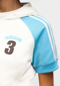 Λευκό αθλητικό φούτερ με μπλε λεπτομέρειες. Διαθέτει τρεις μπλε ρίγες στο κοντό μανίκι, λογότυπο "adidas" και τον αριθμό "3" σε καφέ χρώμα.