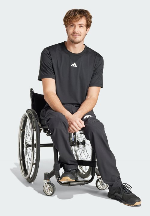 Camiseta deportiva negra con logo blanco, combinada con pantalones oscuros, usada por una persona en silla de ruedas. Zapatillas deportivas negras.