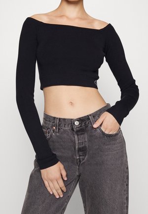 Kobieta ubrana w czarny crop top z długim rękawem odsłaniający ramiona oraz wyblakłe czarne dżinsy z wysokim stanem, z jedną ręką w kieszeni.