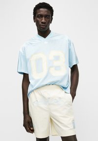 Φωτεινό γαλάζιο δίχτυ με λαιμόκοψη σε σχήμα V και μεγάλο κίτρινο τυπωμένο "03". Συνδυάζεται με κρεμ σορτς που έχουν κεντημένο το "adidas" και ένα έμβλημα.