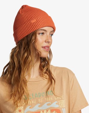 Berretto a maglia a coste arancione con una vestibilità aderente. Presenta un discreto patch con il logo. Indossato con una t-shirt grafica beige che mostra motivi colorati.