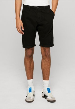 Mand iført sorte shorts med opsmøgede kanter, hvide sokker og hvide sneakers med sorte striber, står med hænderne i lommerne.