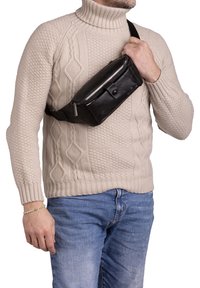 Marsupio in pelle nera con chiusura zip, dettaglio con bottone frontale e cintura regolabile, indossato sopra un pull beige chiaro.