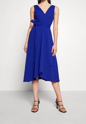 Femme portant une robe portefeuille sans manches bleu roi avec un col en V et un ourlet asymétrique, assortie à des talons hauts noirs à brides.