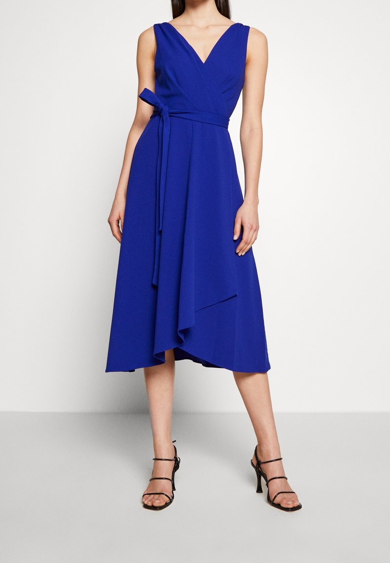 Femme portant une robe portefeuille sans manches bleu roi avec un col en V et un ourlet asymétrique, assortie à des talons hauts noirs à brides.
