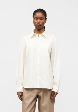 Γυναίκα με πλεξούδες που φοράει λευκό πουκάμισο με κουμπιά και μπεζ παντελόνι, στέκεται μπροστά σε μια απλή λευκή επιφάνεια, κοιτάζοντας προς τα εμπρός.