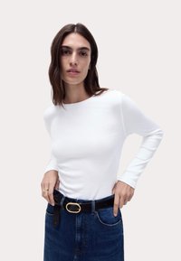 Weißes langärmliges Rippoberteil, kombiniert mit einem dunklen Jeansrock. Das Modell trägt einen schwarzen Gürtel mit einer goldenen ovalen Schnalle.