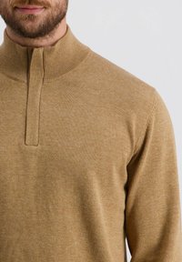 Tan gebreide trui met een hoge kraag en een half-rits ontwerp. Het materiaal lijkt zacht met een gladde textuur en een subtiel gemêleerd patroon.