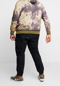 Mikina s kapucí s hnědým a béžovým tie-dye vzorem, zelenými akcenty, doplněná černými zúženými kalhotami a zelenými teniskami.