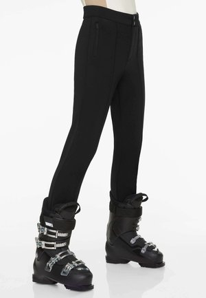 Pantalons de ski noirs au design ajusté, avec des poches zippées et une texture lisse. Associés à des chaussures de ski noires avec des boucles en métal.