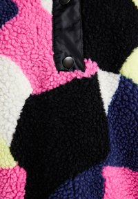 Farverigt sherpa-stof med sorte, pink, hvide, marineblå og gule hexagonale mønstre, med glatte sorte akcenter og trykknapper.