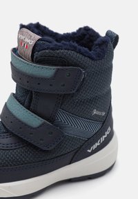 Jūras zila potīšu zābaku ar teksturētu audumu, pelēkiem siksnām un mīkstu vilnas oderi. Iezīmēts ar ūdensnecaurlaidīgu Gore-Tex zīmolu un zigzaga rakstu.