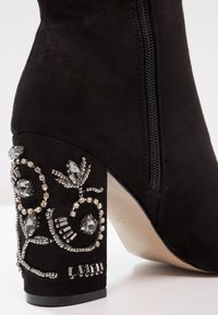Bottine en daim noir avec fermeture éclair et talon carré orné de perles et de cristaux formant des motifs floraux.