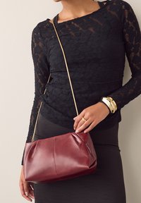 Bordeauxrode leren handtas met een gladde textuur, voorzien van een gouden kettingriem en een gevouwen ontwerp, vastgehouden door een persoon in een zwarte kanten top.