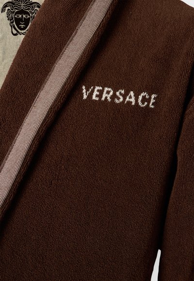 Σκούρο καφέ υφασμάτινη υφή με κέντημα λογότυπου Versace σε λευκό και μπεζ διακοσμητική περίμετρο με μοτίβο που περιλαμβάνει μαύρο έμβλημα κεφαλής Μέδουσας.