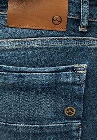 Κοντινή εικόνα μπλε τζιν από τζην ύφασμα με καφέ δερμάτινο έμπλαστρο και μικρό κυκλικό λογότυπο ραμμένο κοντά στην πίσω τσέπη.