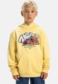 Młody chłopiec z blond włosami noszący żółtą bluz�ę z kapturem z grafiką czerwonego samochodu i niebieskie spodnie, stojący na tle białym.