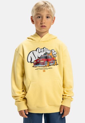 Junger Junge mit blonden Haaren, der einen gelben Kapuzenpullover mit rotem Auto-Design und blauen Hosen trägt, vor einem weißen Hintergrund.