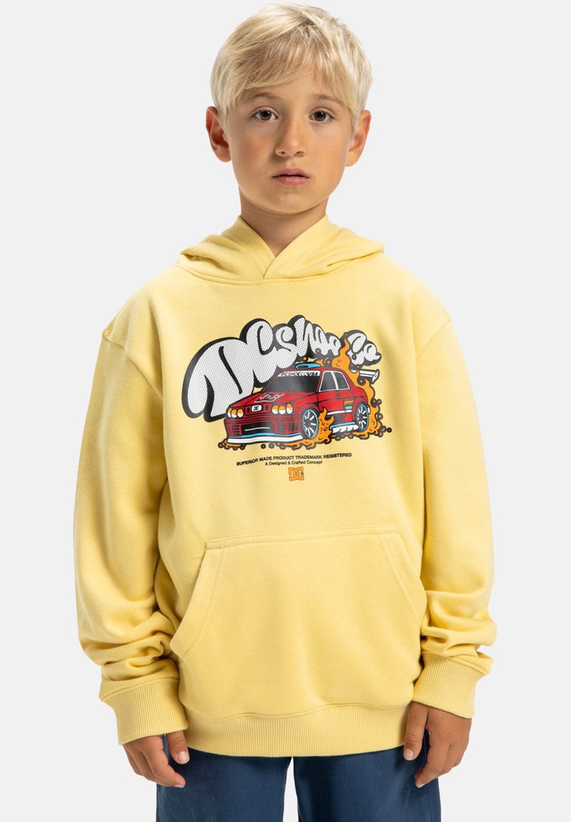 Młody chłopiec z blond włosami noszący żółtą bluzę z kapturem z grafiką czerwonego samochodu i niebieskie spodnie, stojący na tle białym.