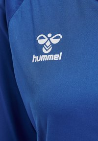 Blaues Sporthemd mit glatter Textur und einem weißen Hummel-Logo. Das Design hat einen einfachen, modernen Schnitt.