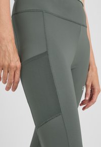 Hoggetaille sportlegging in olijfgroen, met gaaspanelen, stikdetails en een zak op de dij voor gemakken.