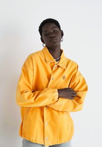 Gul denimjakke med krage, trykknapper, elastiske mansjetter og logo på brystet. Modellen poserer med armene krysset foran en hvit bakgrunn.