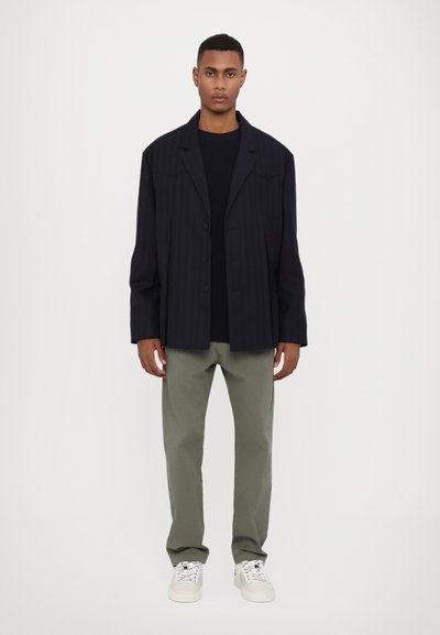 Marineblau gestreifter, übergroßer Blazer mit Reverskragen und Knopfleiste, kombiniert mit einem schwarzen Pullover und olivgrünen Hosen. Weiße Sneaker.