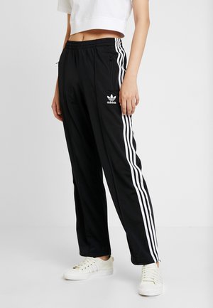 Persona con pantalones deportivos negros de Adidas con rayas blancas y zapatillas blancas, de pie contra un fondo claro y liso.