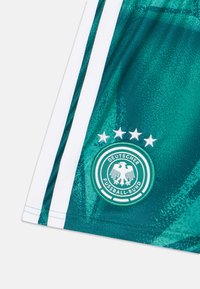 Zaļas futbola šorti ar baltām sānu svītrām, uz kuriem redzams Deutscher Fussball-Bund (Vācijas Futbola Savienības) logotips un virs tā četras zvaigznes. Auduma tekstūra.