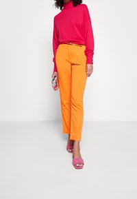 Różowy sweter z golfem, pomarańczowe eleganckie spodnie i różowe sandały na obcasie. Trzyma torbę kopertową w wielokolorowy wzór. Gładkie tekstury materiałów.