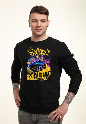 Mand står med hænderne på hofterne iført sort sweatshirt med farverigt graffiti-design af en blå bil og teksten "NEW STANDARD."
