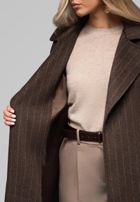 Abrigo de lana a rayas marrones, suéter de punto beige y pantalones ligeros beige. Cinturón marrón y uñas bien cuidadas. El forro es de un tono más claro.