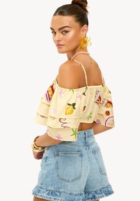 Blouse jaune à épaules dénudées avec motifs colorés de fruits et de boissons, assortie à un short en denim bleu avec ourlets à volants. Accessoires dorés visibles.