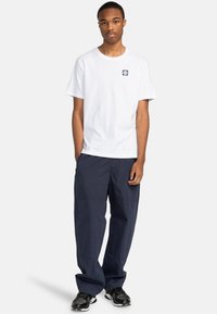 Camiseta de algodón blanca con un pequeño parche de logo en navy, combinada con pantalones sueltos de navy. El modelo lleva unas zapatillas negras con un diseño texturizado.