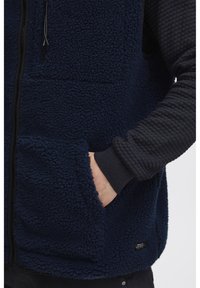 Mornarska flis prsluk s mekim, teksturiranim finišom, zatvorenim prednjim dijelom s patentnim zatvaračem, dva bočna džepa i rebrastim sivim rukavima. Diskretna oznaka brenda na rubu.