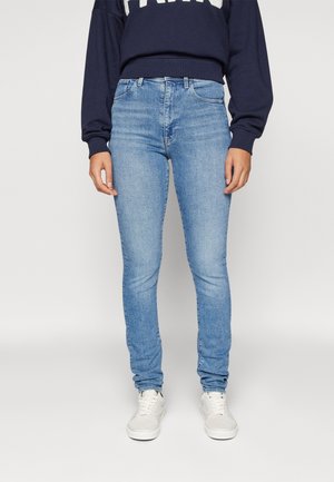 Personne portant un jean slim bleu clair, des baskets blanches et un sweat court bleu marine avec des lettres blanches, debout devant un fond uni.