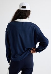 Dunkelblaues Sweatshirt mit einem weißen Kragen, mit lockerer Passform und abgesenkten Schultern, kombiniert mit dunkelblauen Leggings mit weißen Streifen.