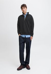 Noor mees, kes kannab musta poollukuga pusa sinise nööbitava särgi, tumedate teksapükste ja pruuni nahkjalanõudega, seisab käed taskutes.