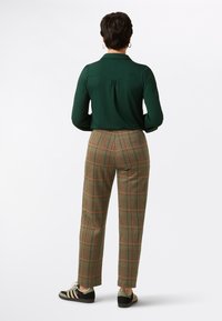 Groene blouse met stippen en lange mouwen, gecombineerd met bruine broek met ruitpatroon en oranje accenten, van achteren weergegeven. Zwarte sneakers.