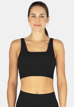 Frau mit zurückgebundenen Haaren, die einen schwarzen Sport-BH mit quadratischem Ausschnitt und schwarze Leggings mit hohem Bund trägt, steht vor einem schlichten Hintergrund.