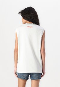 Kreklu bez piedurknēm, balts, izgatavots no kokvilnas, ar oranžu "Wrangler" logotipu aizmugurē. Tam ir brīva piegriezuma forma un gluda tekstūra. Valkā ar džinsu šortiem.