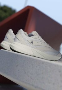 Paire de baskets athlétiques blanc cassé avec mesh texturé et panneaux lisses reposant sur une surface en béton, inclinée avec une structure géométrique rouge floue en arrière-plan.