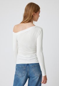 Witte lange mouwen top met een off-shoulder ontwerp, voorzien van een gerimpelde textuur aan de achterkant en gecombineerd met blauwe denim jeans.