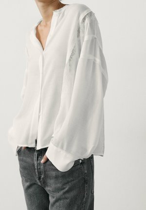 Personne portant un chemisier blanc translucide à manches longues bouffantes et un jean gris foncé, mains dans les poches, sur un fond clair uni.